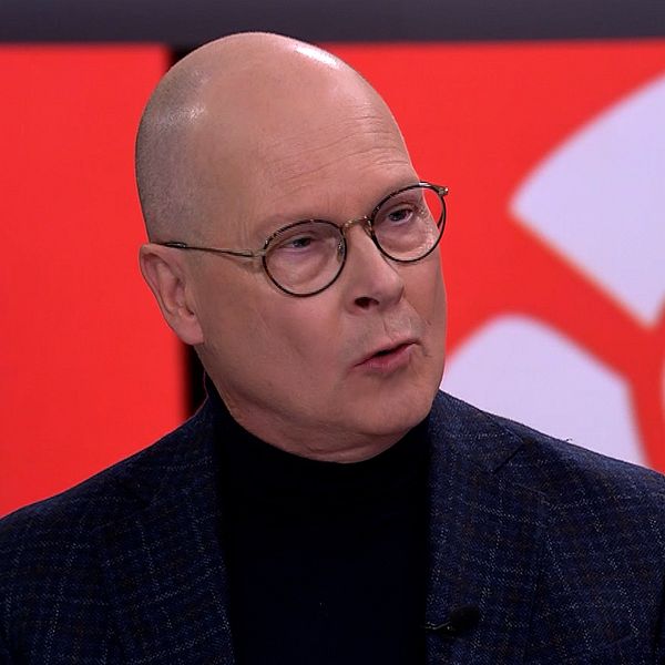 Mats Knutson i kostym står framför en färgglad bakgrund med röda och vita mönster, vilket indikerar en tv-studio eller liknande miljö. Personen verkar vara involverad i en diskussion eller intervju.