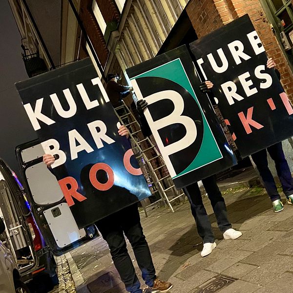 Tre personer håller upp stora skyltar som visar texten ”KULTUR”, ”BAR”, ”ROCK” och ”B” i olika färger och storlekar. Bakgrunden är en stadsmiljö på kvällen, med låg belysning och några byggnader synliga.
