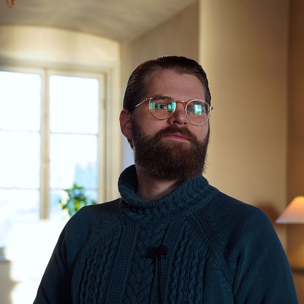 Cristoffer Tidelius, en man med skägg och glasögon, står i ett slottsrum och tittar in i kameran.