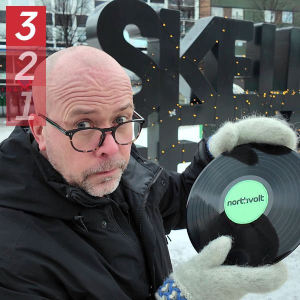 Reporter Patrik Samuelsson i vinterkläder håller upp en svart vinylskiva med etiketten ”Northvolt” framför en stor dekorativ skylt med bokstäverna ”SKELLEFTEÅ” och en ljussatt sfärisk installation i bakgrunden. Det är vinter och omgivningen är snöklädd.