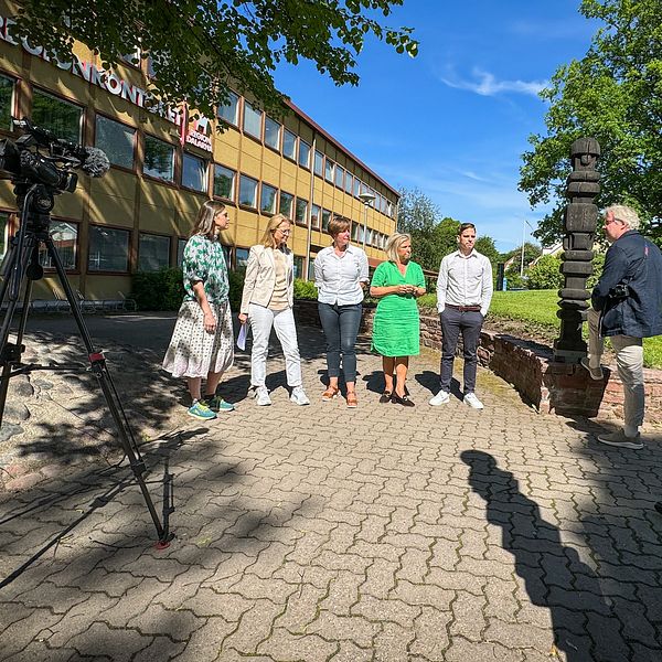 En grupp människor står i solskenet framför en stor gul byggnad. En filmkamera är riktad mot dem och flera journalister lyssnar på vad de säger.