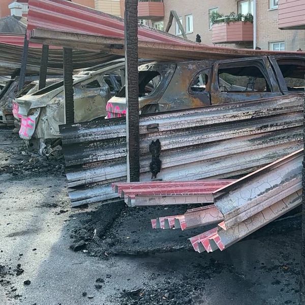 Skadade bilar parkerade under en delvis förstörd carport, med brända och lutande takplåtar. Markytan visar tydliga tecken på brand och förstörelse. I bakgrunden syns en byggnad med lägenheter.