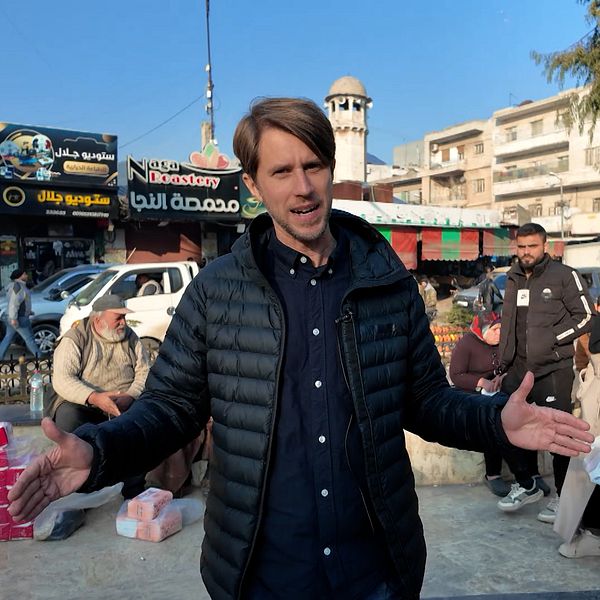 Tomas Thorén står med utsträckta armar på en livlig marknadsplats, omgiven av människor som bär på kassar. I bakgrunden syns affärer och ett minaret.