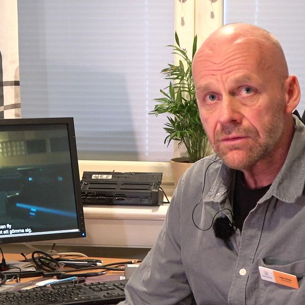 En person sitter vid ett skrivbord med en datorskärm i bakgrunden. Skärmen visar en scen från med någon som går i en skolaula. Ett skrivbord är belamrat med papper och kontorsutrustning, och det finns en grön växt i fönstret bakom.