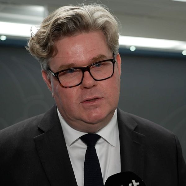 En person i kostym står framför en vägg med en subtil design, han ser ut att pratat i en mikrofon.