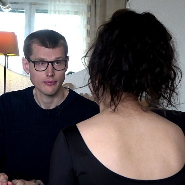 En kvinna sitter vänd mot en man i en inomhusmiljö. Kvinnan har svart, axellångt hår och bär en svart klänning med öppen rygg. Mannen är klädd i en mörk tröja och tittar mot kvinnan. I bakgrunden syns en stor spegel samt en lampa. Rummet verkar vara avskalat och stilrent.