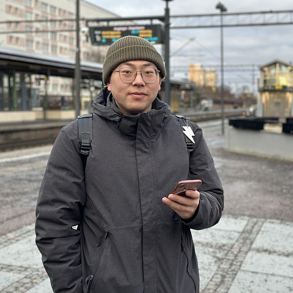 Person i mörk jacka står på en tågperrong och håller en smartphone.