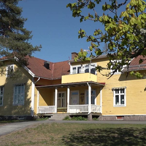 Gul trävilla med rött tak och veranda, omgiven av träd under klarblå himmel.