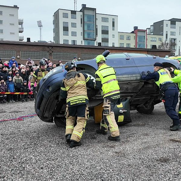 Brandmän och räddningspersonal välter en bil på en grusplan inför en publik som står bakom avspärrningar. Flera åskådare har samlats för att titta på demonstrationen, och moderna lägenhetsbyggnader syns i bakgrunden.
