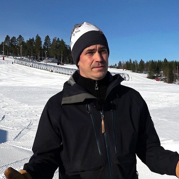 Person i skidjacka och mössa står på snötäckt skidanläggning med klarblå himmel.