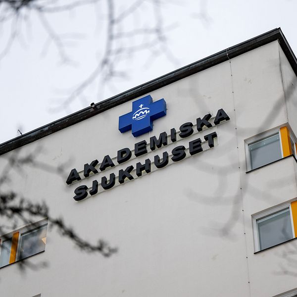Klocka på Akademiska sjukhuset med tydlig logotyp och namnskylt på fasaden, omgiven av trädgrenar.