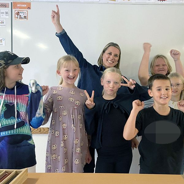 Grupp av barn som poserar framför en whiteboard, glada och applåderande. Några har händerna upp i luften och visar tecken på seger. Bakom dem hänger två gyllene stjärnor dekorationer. Klassen verkar vara i en skolmiljö.