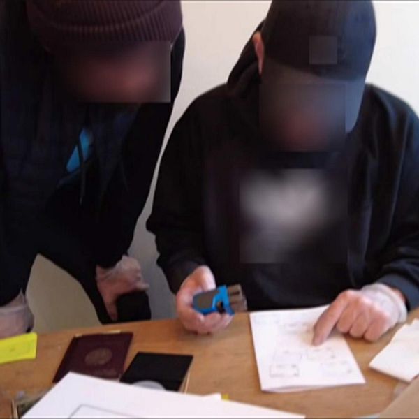 Två män granskar id-handlingar vid ett bord, ansiktena är suddade för anonymitet.