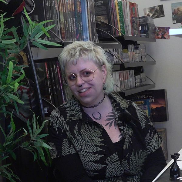 Lovisa Falk sitter bland spel och böcker i spelbutiken i Falun. Hon ha blont hår, glasögon och en bladmönstrad skjorta.