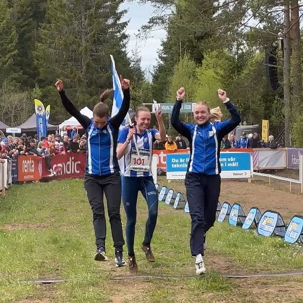 Tre idrottare firar med armarna i luften vid en mållinje, publik hejar längs sidorna.