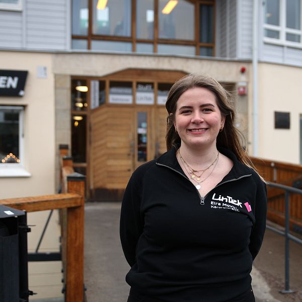 En kvinna står framför byggnaden med namnet ”Kårallen” synlig på fasaden. Hon bär en svart tröja med en logotyp. I bakgrunden syns en träbro som leder in i byggnaden, samt fönster och en del av den omgivande miljön.