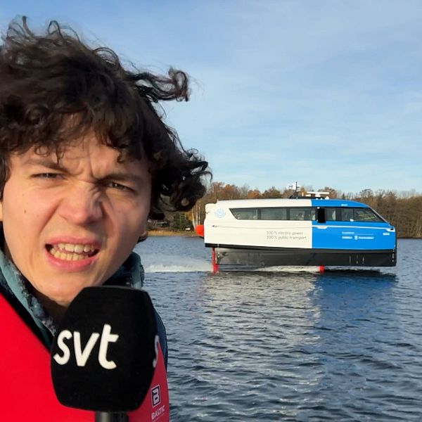 En person i en röd flytväst står vid vattenkanten med en mikrofon i handen. I bakgrunden syns en innovativ vattenfarkost som åker fram på vattnet, omgiven av en naturskön miljö med träd och en klar himmel.