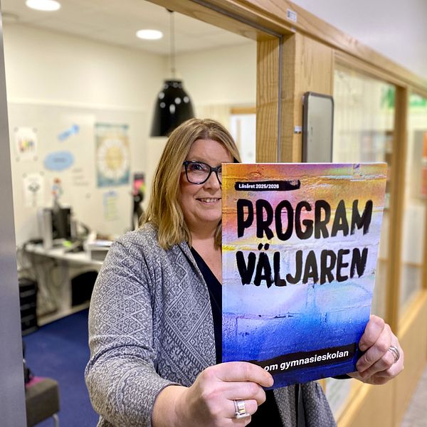 En person står i en korridor och håller upp en färgglad broschyr med texten ”Program Väljaren” som verkar handla om gymnasieskolan. I bakgrunden syns ett kontor och en korridor med färgglad dekor.