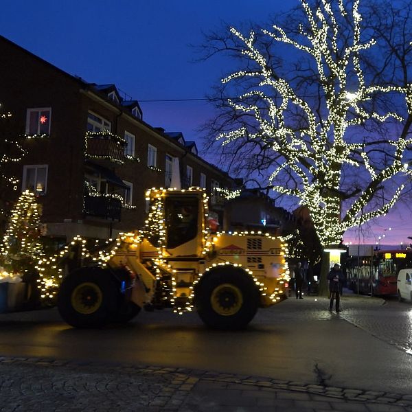 En belyst hög lastmaskin pryds av juldekorationer och kör genom en stadsgata som är vackert utsmyckad med ljus. I bakgrunden syns ett stort försedd träd även det täckt med ljus, samt flera byggnader med julprydnader. Skymningen ger en stämningsfull atmosfär i bilden.