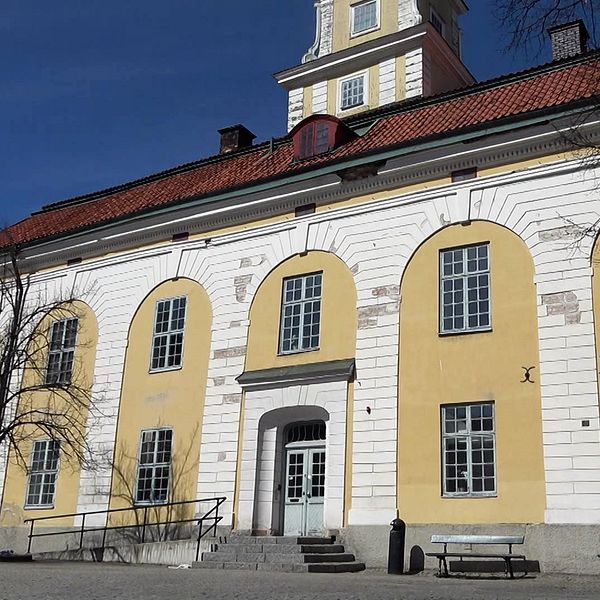 Gul och vit byggnad med flera fönster, blå himmel och några träd i förgrunden. Fasaden är skadad.
