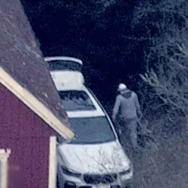 En man i grå träningskläder står vid en vit bil som är parkerad vid skogskanten. I bakgrunden syns ett rödaktigt hus med vita fönster. Området är omgiven av tät vegetation.