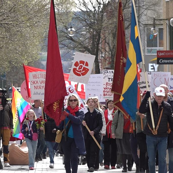 Demonstrationståg med svenska flaggan och röda fanor, människor bär plakat och skyltar.