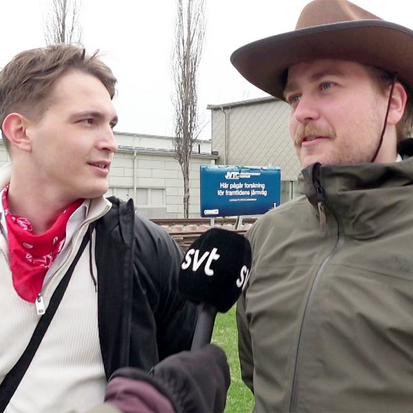 Två personer intervjuas utanför en byggnad. En av dem har en cowboyhatt på huvudet.