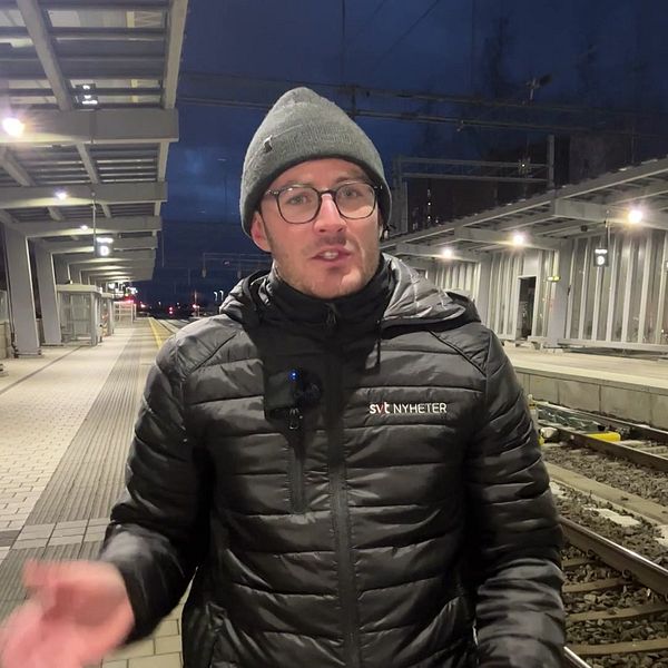 En person står på en tågstation under kvällstid, iförd en svart jacka och en mössa. Stationen har en modern arkitektur med lyktor som skapar en belyst miljö. Tågrälsen syns i bakgrunden tillsammans med stationens plattform.