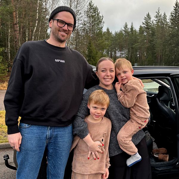 En familj står utomhus vid en bil, omgivna av träd och en vägskylt i bakgrunden. Två barn står framför en vuxen, som håller ett av barnen i famnen, medan det andra barnet står bredvid. De är klädda i avslappnade kläder och ler mot kameran.
