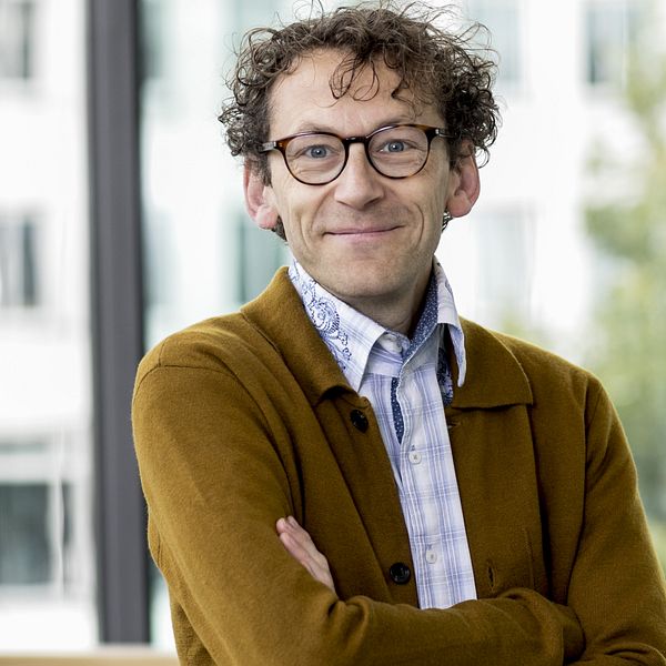 En person, Åke Ingerman, med korslagda armar står i ett ljust rum med stora fönster som släpper in naturligt ljus. Personen har på sig en brun kofta över en mönstrad skjorta. I bakgrunden syns oskarpa kontorsmiljöer.