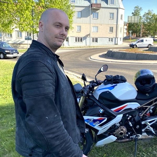 Johan Andersson i MC-jacka står framför sin motorcykel som står i gräset vid en rondell