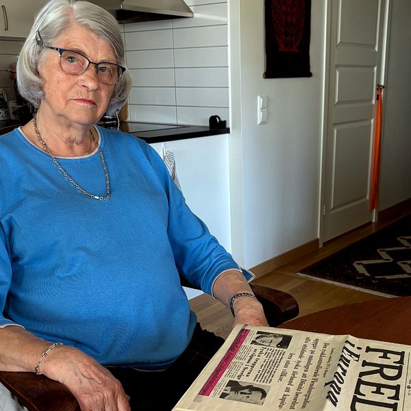 Äldre person i blå tröja sitter vid köksbord och läser en tidning med rubriken ”Fred”.