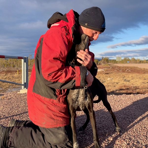 Man i röd jacka kramar en hund på grusväg under blå himmel.