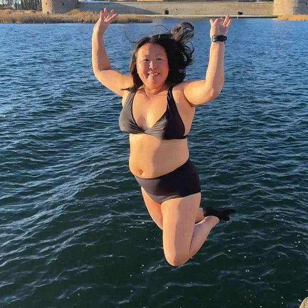 En person är i luften på väg att hoppa i vattnet från en brygga, iförd en bikini. Vattnet är klart och reflekterar solljuset, och i bakgrunden syns Kalmar slott. Det är en solig dag, vilket skapar en livlig och energisk atmosfär.
