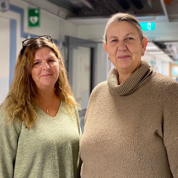 Två personer står i en sjukhuskorridor, klädda i komfortabla tröjor. Korridoren har flera dörrar och släpljus som ger en lugn atmosfär. Bakgrunden är suddig men visar att det är en vårdmiljö.