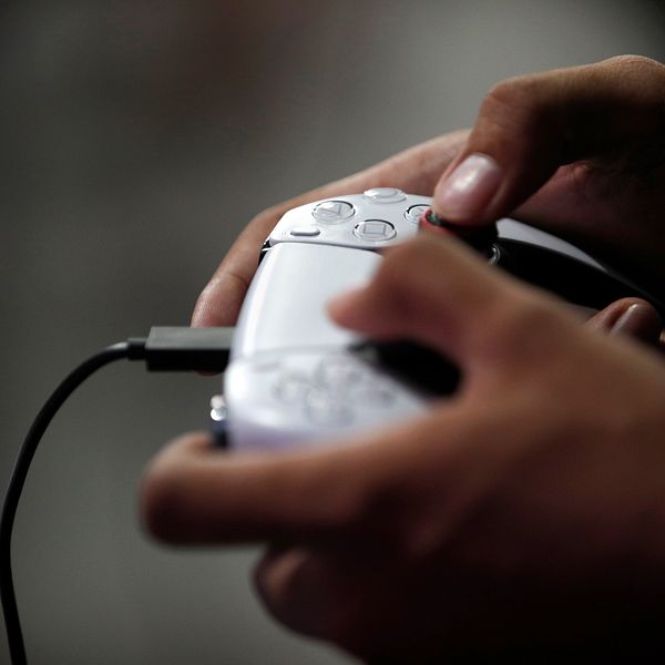 Händer som håller en vit spelkontroll, troligtvis en PlayStation 5-kontroll, i en mörk bakgrund.