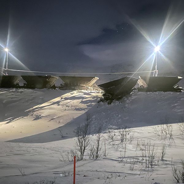 strålkastare över malmvagnar på vall, snötäckt mark. En vagn hänger ner längs vallen