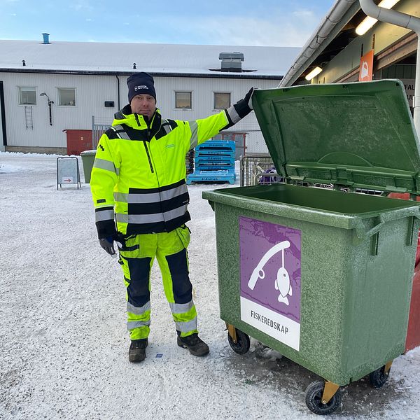 En person i en reflexjacka står vid en öppen grön avfallscontainer märkta med texten ”Fiskeredskap” på en vintrig plats med snö. Bakgrunden visar en del av en byggnad och andra containrar i närheten.