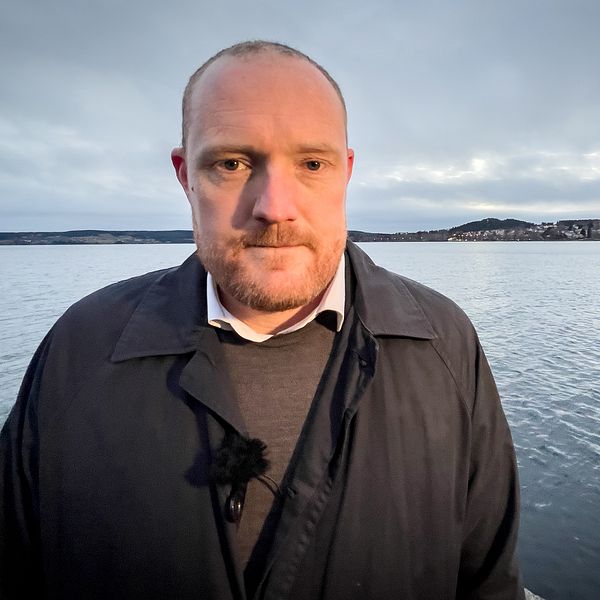 En person står vid vattnet med en sjö och en strandlinje i bakgrunden. Himlen är mulen och skapar en dämpad atmosfär. Personen är klädd i en mörk kappa och har ett mikrofonfäste på bröstet.