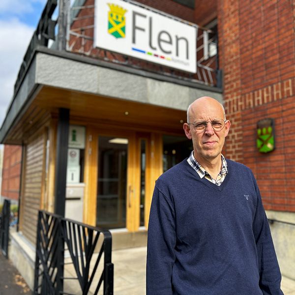 En person står framför entrén till ett byggnad med en skylt som visar namnet ”Flen”. Byggnaden har trä- och tegelarkitektur, och det finns en grön emblem på väggen. Det är en klar dag med lite moln på himlen.