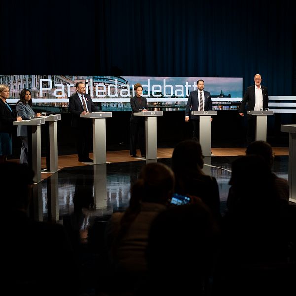 Partiledardebatt på scenen, partiledare står bakom varsitt podium, publik i förgrunden.