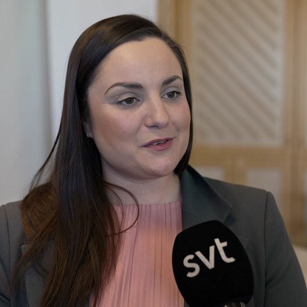 Kvinna i rosa tröja med SVT-mikrofon, stol och bord i bakgrunden.