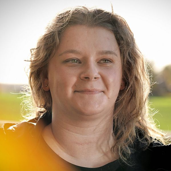En person i profil står i en naturskön utomhusmiljö med grönska i bakgrunden. Ljusa färger och mjukt ljus skapar en varm stämning.
