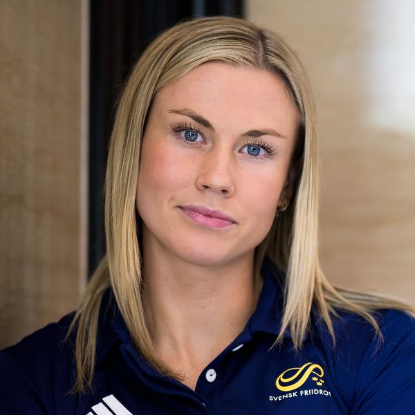 Person i mörkblå sporttröja står lutad mot en ljus vägg, armar i kors.