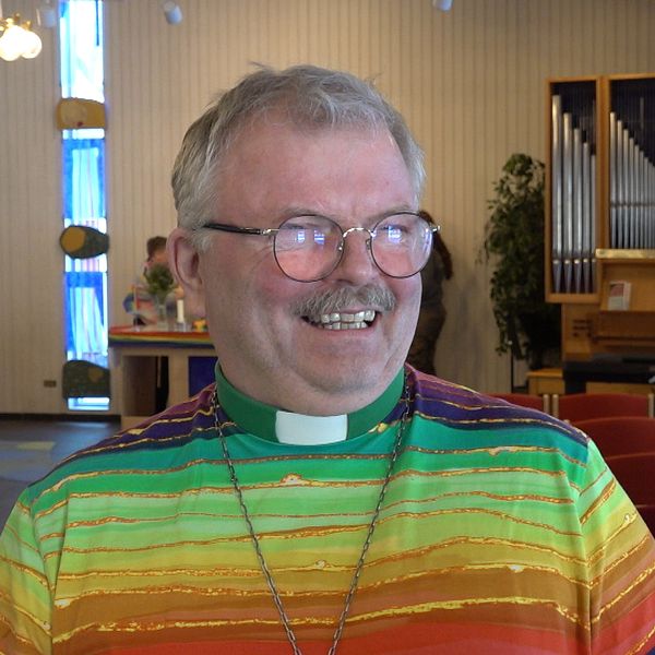 En kyrka med orgel och rader av röda stolar i bakgrunden. En man i regnbågströja ler.
