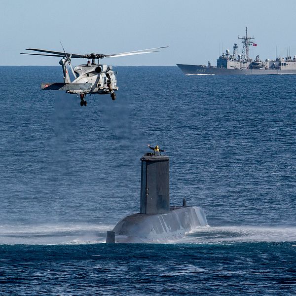 Helikopter ovanför ubåt på havet, krigsfartyg i bakgrunden.