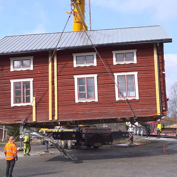 Hembygdsgården i Hackås lyfts av en stor kranbil till en väntande trailer. Byggarbetare i varselkläder övervakar arbetet. Huset är timrat och rött med vita foder runt fönstren.