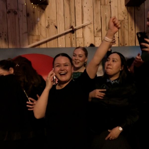En grupp människor firar med höjda händer, stående nära en trävägg.