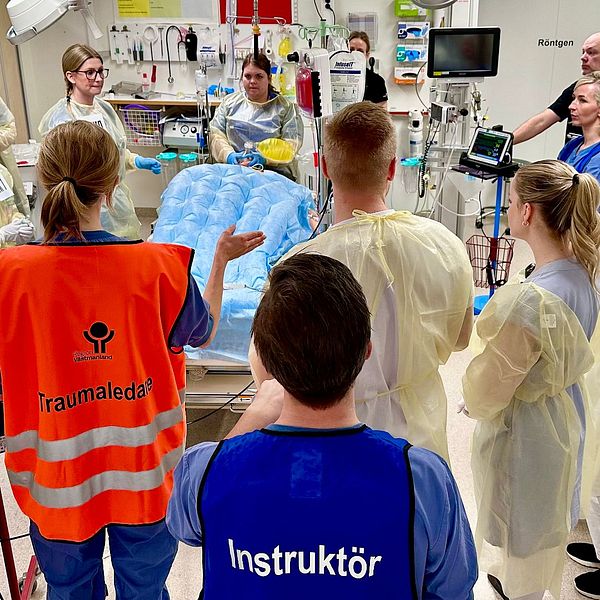 En grupp sjukvårdspersonal i skyddskläder samlas i ett sjukhusrum, ledda av en person i orange väst med texten ”Traumaledare” och en annan i en blå väst med texten ”Instruktör”. De verkar delta i en medicinsk träning eller simulering med medicinsk utrustning och en säng i mitten av rummet.