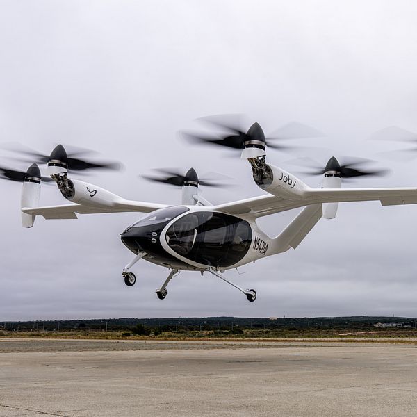 En svartvit elektrisk drönare med flera rotorer som lyfter från en landningsplats. Drönaren har en aerodynamisk design med tydliga propellrar i rörelse, vilket indikerar att den är i flygning. I bakgrunden syns en grönskande och öppen landskap under en övercast himmel.